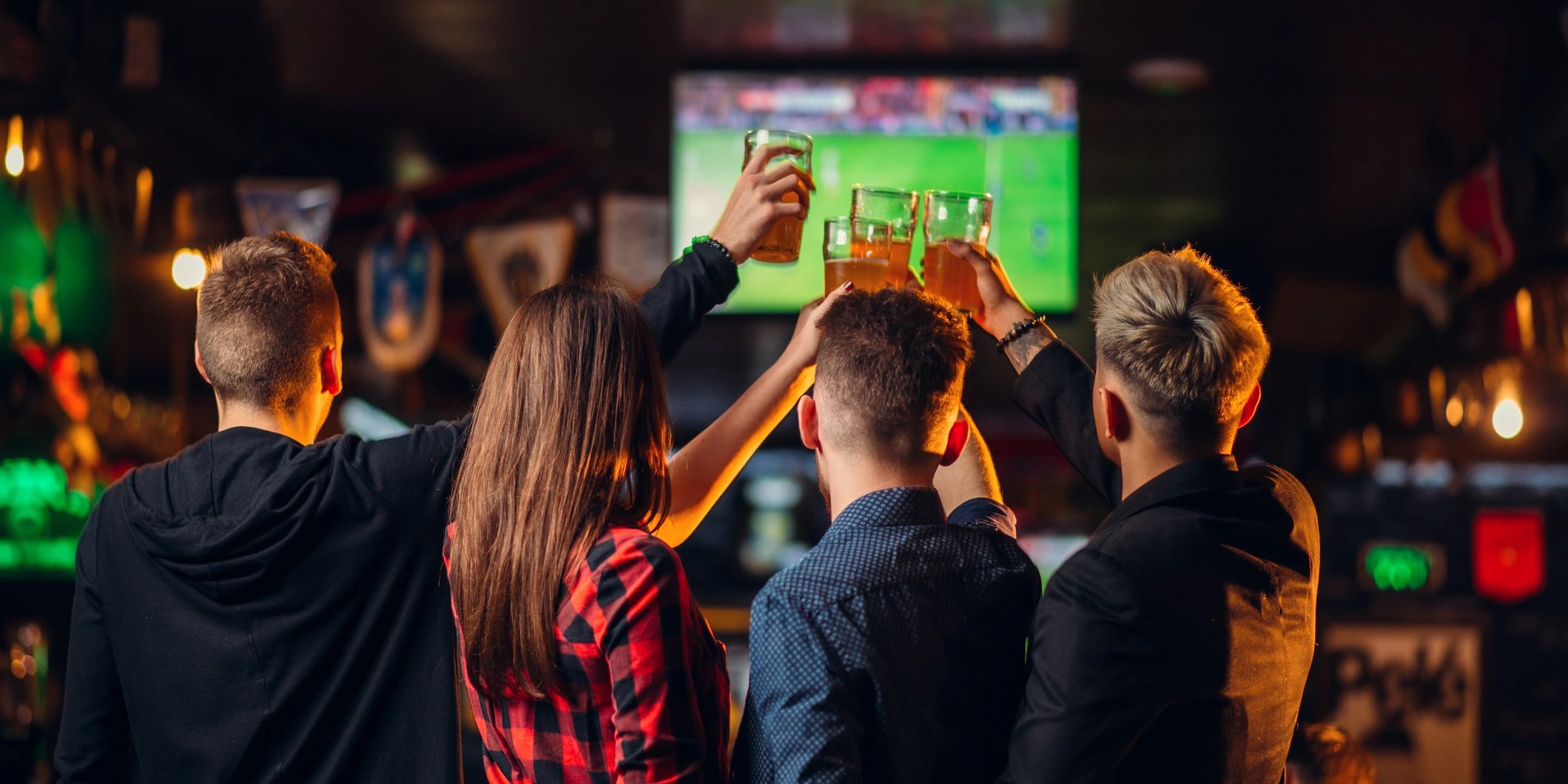 Friends raising beer glasses together while watching a game on the big screen