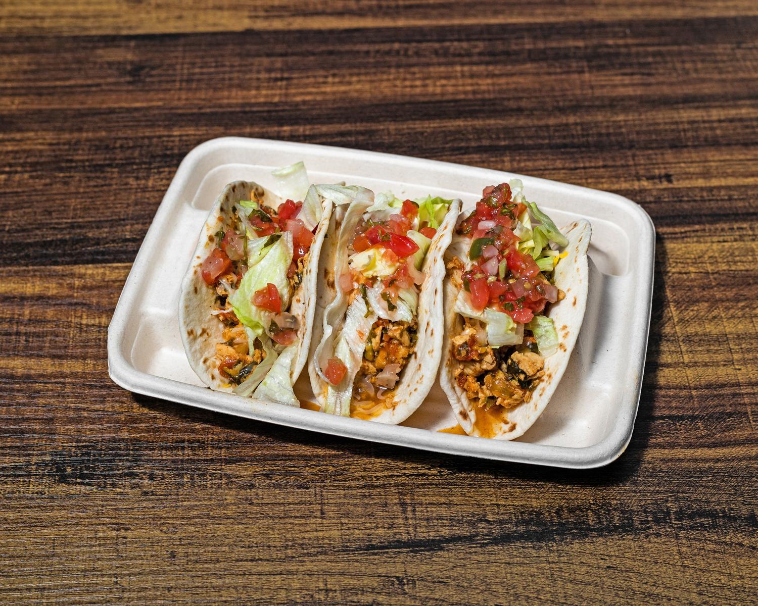 Three street tacos topped with lettuce, cheese and salsa in a cardboard tray