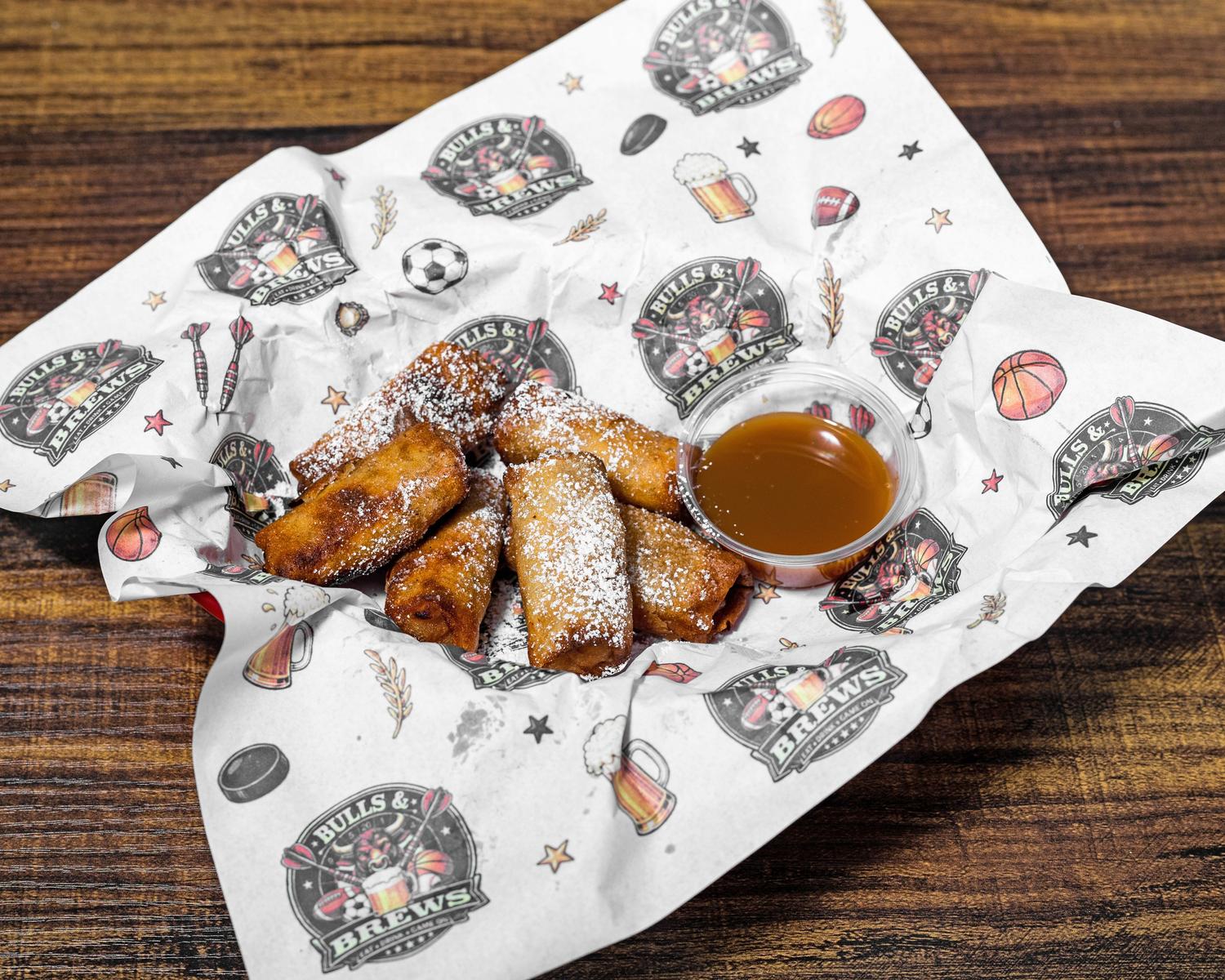Apple pie bites dusted with powdered sugar and served with caramel dipping sauce on Bulls & Brews wrap paper